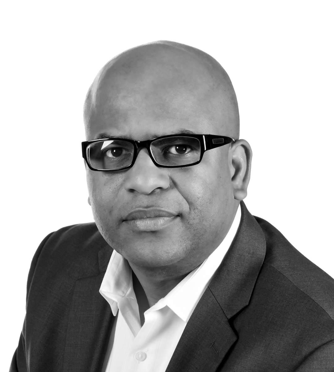 A bald man wearing glasses and a suit is looking at the camera in a black and white photo.