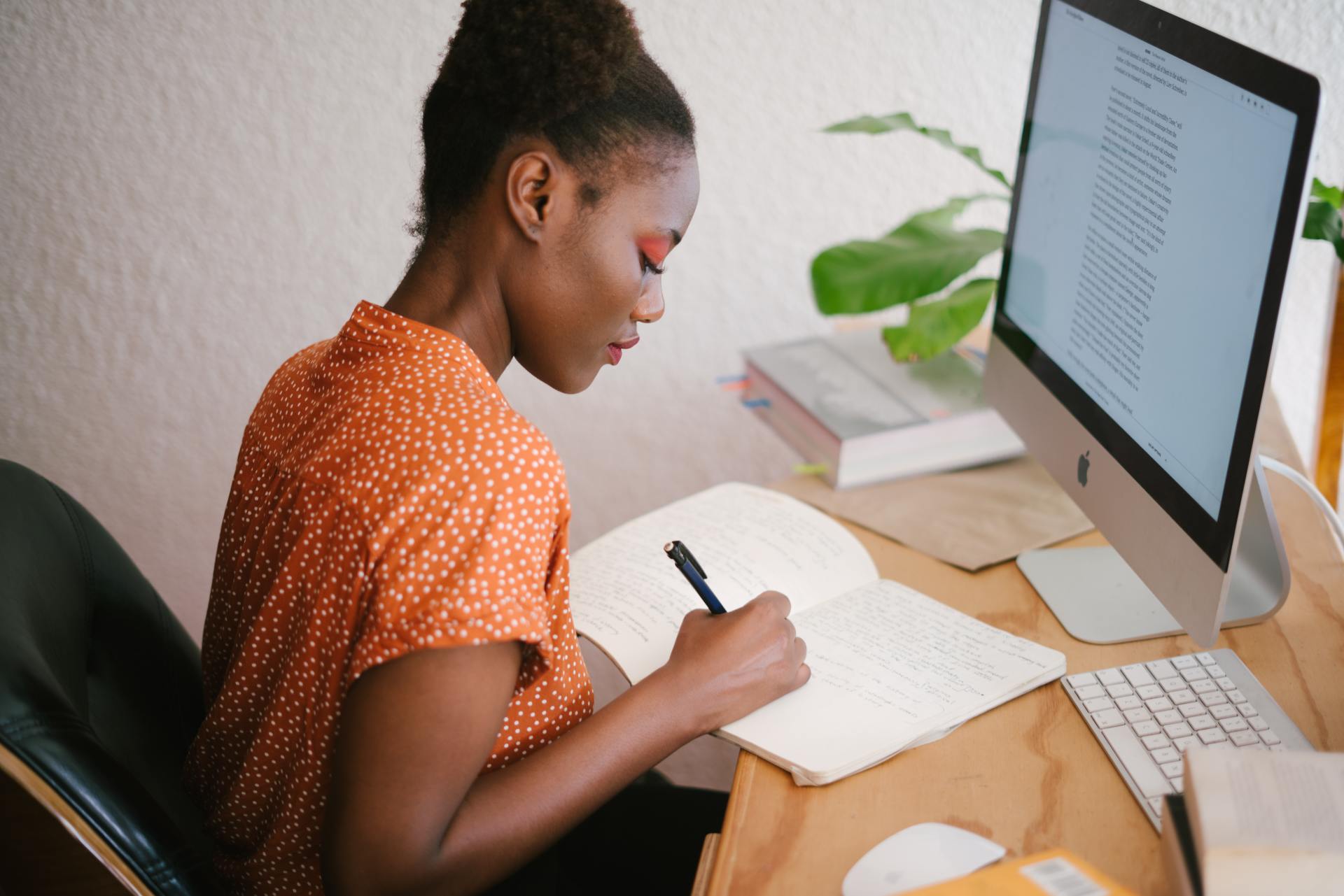 A woman is sitting at a desk with a computer and writing in a notebook.