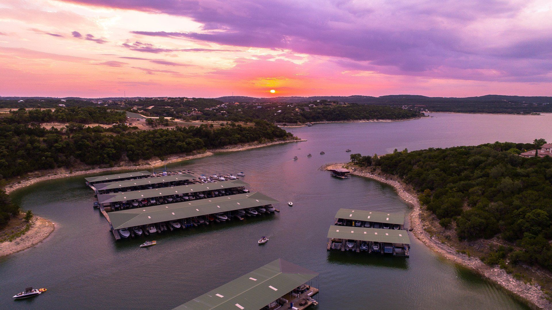 Aerial view of a marina with boat slips on a lake at sunset, with purple and orange skies over forested shorelines.
