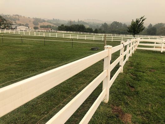 White ranch fence bordering a green pasture with hazy trees and hills in the background