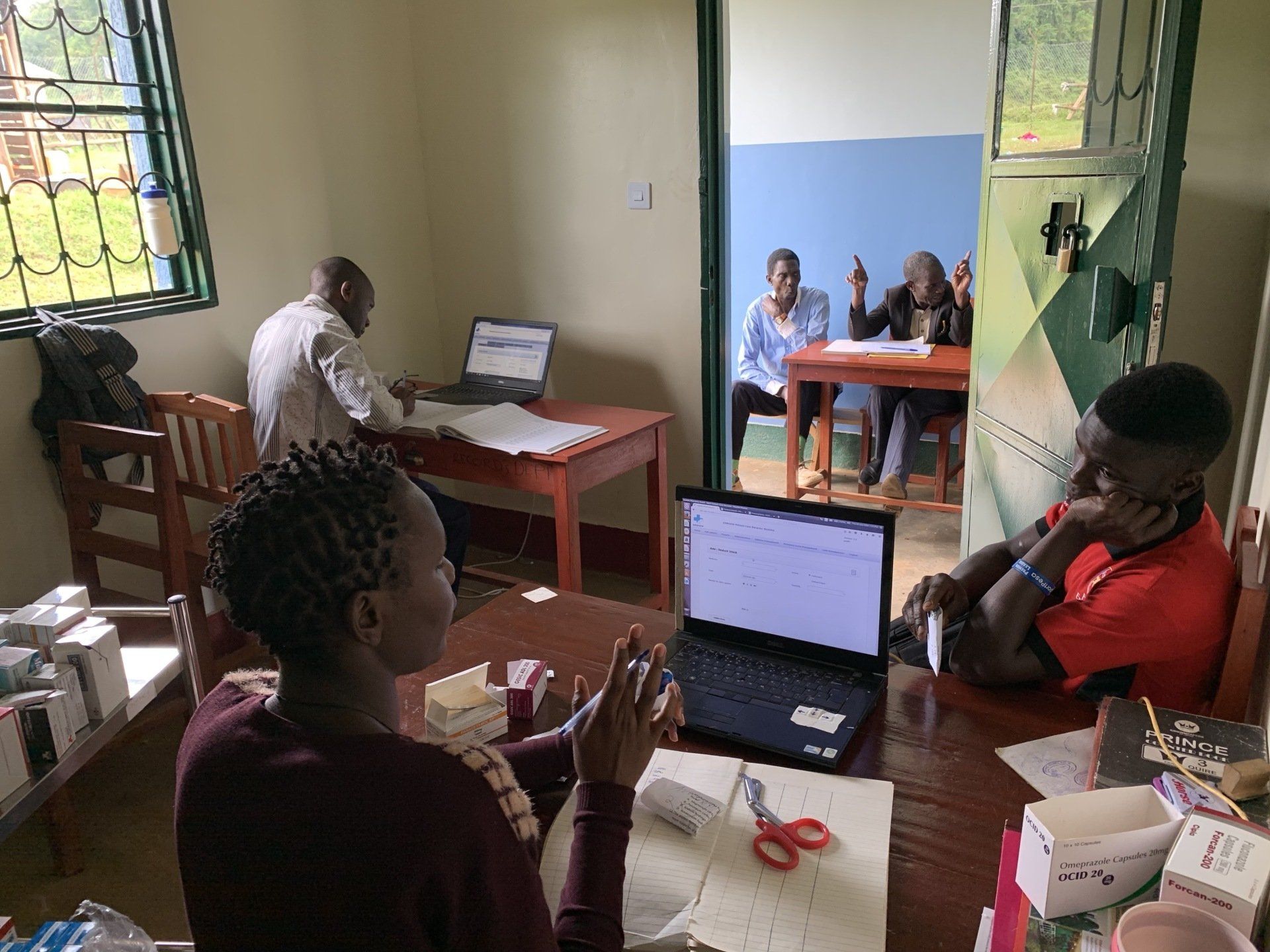 People in a room working on laptops. One woman gestures, others seated at tables.