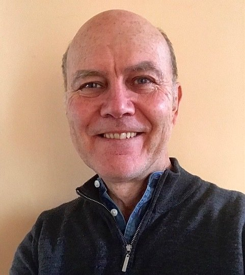 Bald man smiling, wearing a dark zip-up over a blue shirt, against a plain beige wall.