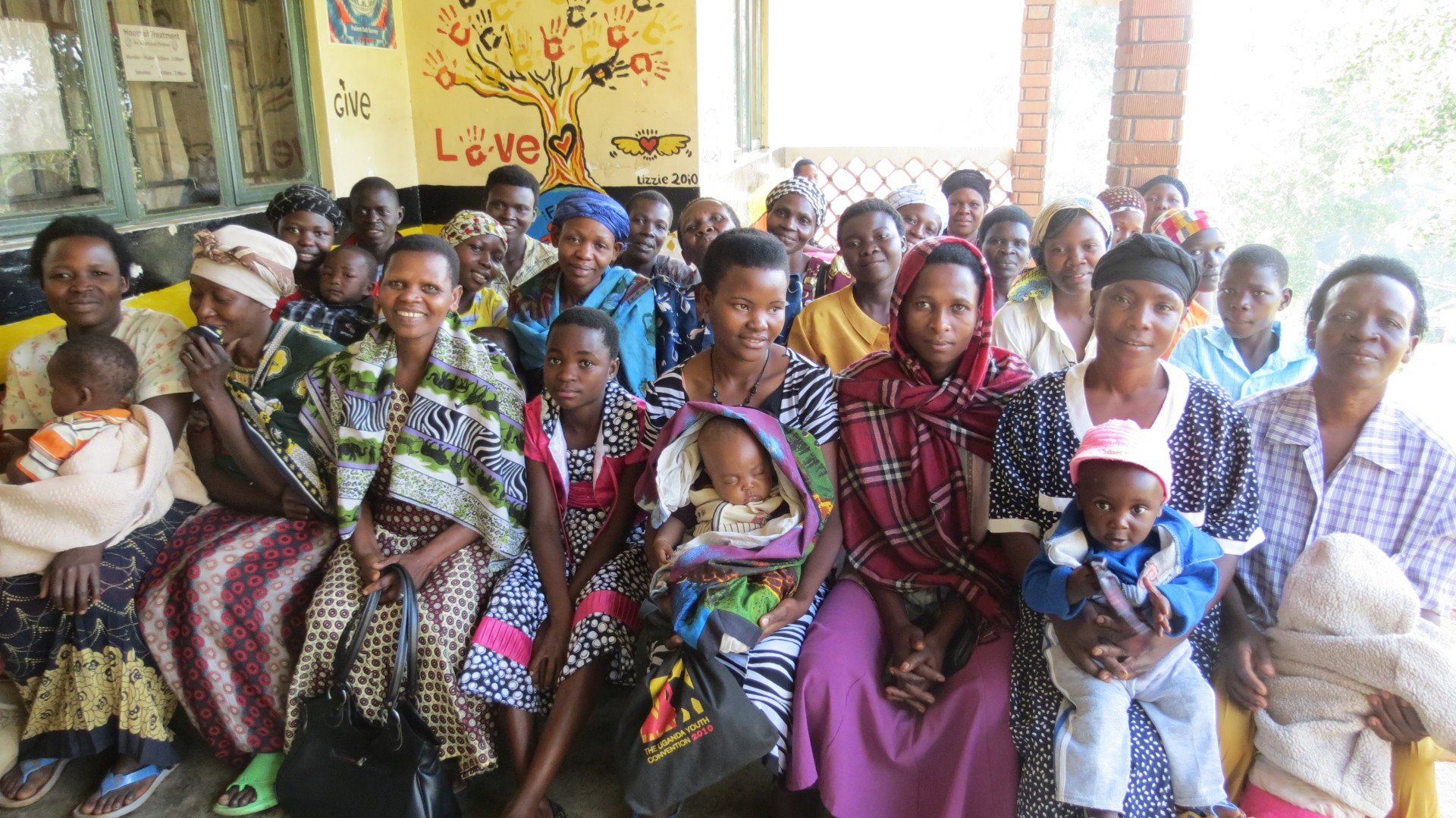 Group of women with babies sitting outdoors, smiling. Yellow and red painted wall background.