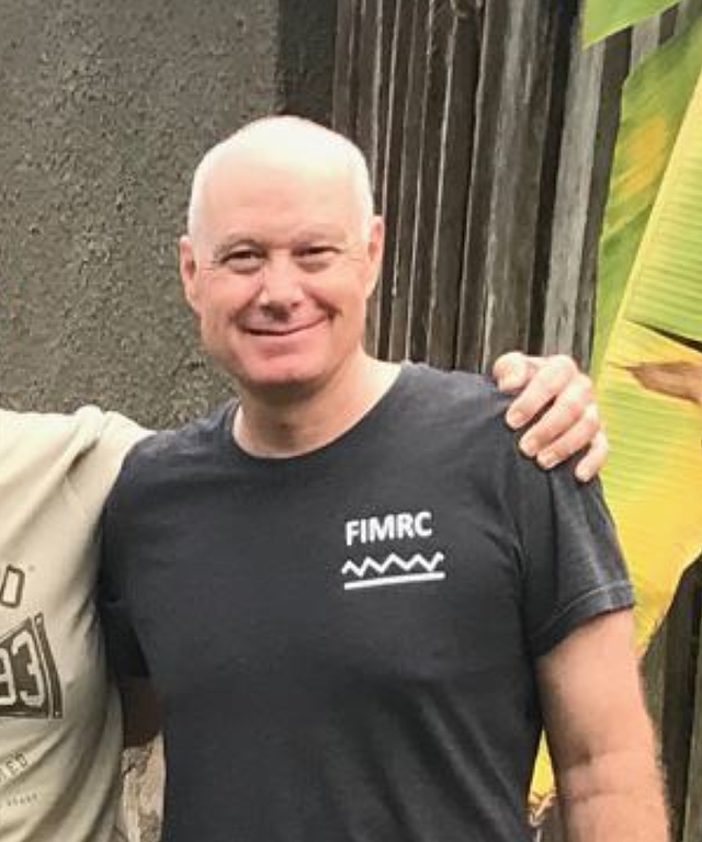 Middle-aged man in black t-shirt with FIMRC logo smiles, arm around someone out of frame, near wood and plants.