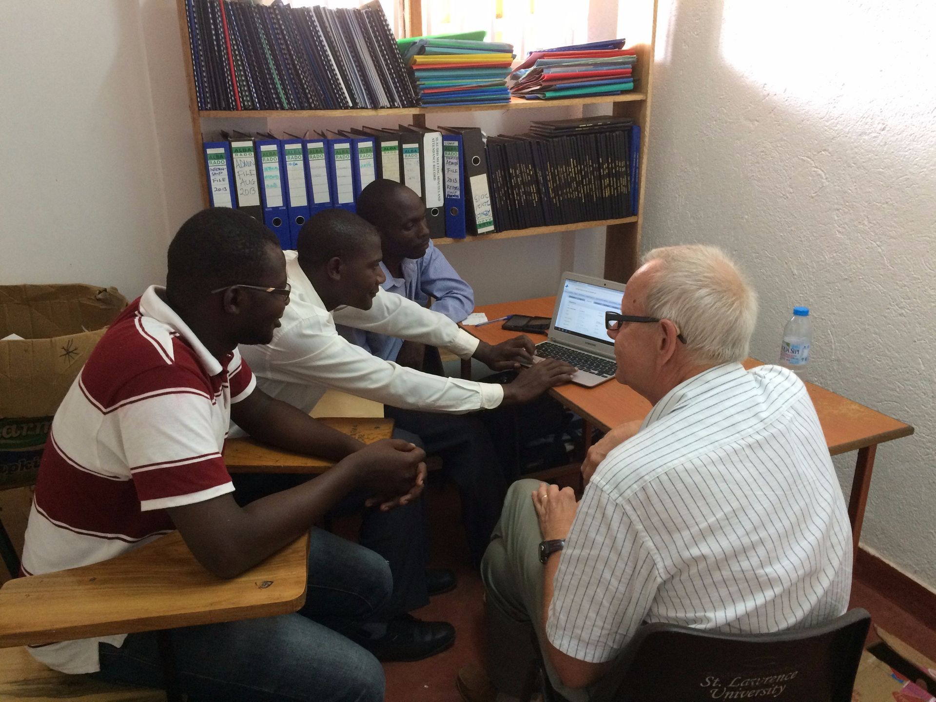 Four men in a small office looking at a laptop. Bookshelves in the background.