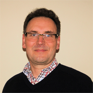 Man wearing glasses, black sweater, checkered shirt, smiles in front of a beige wall.
