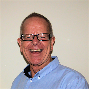 Man with glasses smiles broadly, wearing a blue shirt against a white wall.