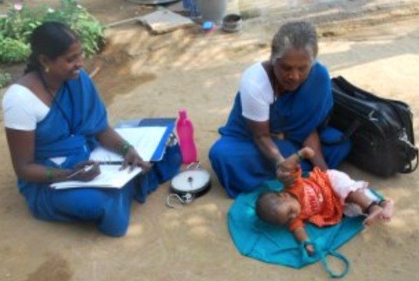 Two women in blue saris weigh an infant outdoors. One records data while the other holds the baby.