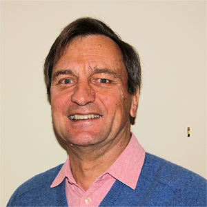 Smiling older man with brown hair, wearing blue sweater over pink shirt, against a white wall.