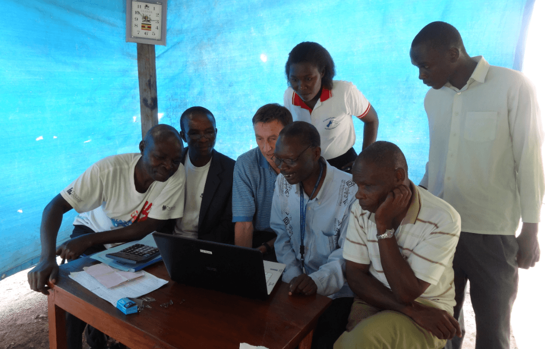 Group of people looking at a laptop screen together outdoors under a blue tarp.