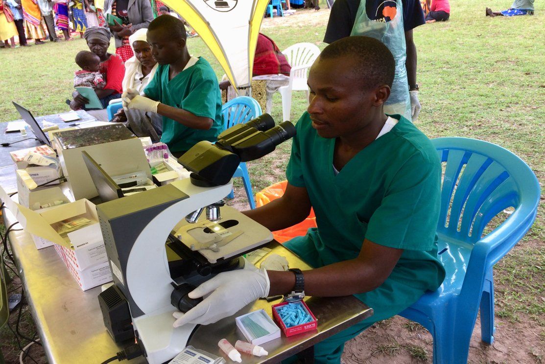 A man in green scrubs uses a microscope at an outdoor medical clinic; others assist in the background.