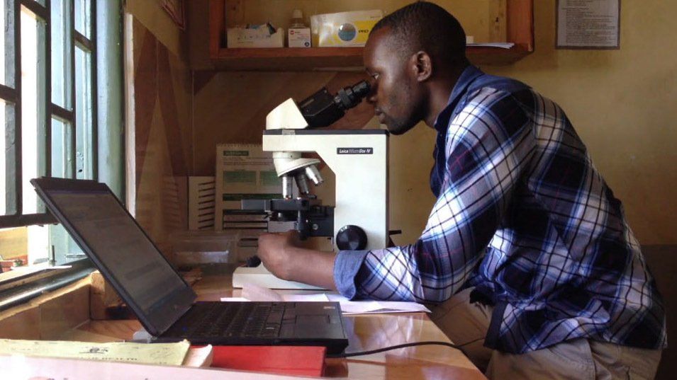 Man in plaid shirt looking into a microscope, with a laptop open on a desk beside him. Indoor setting.