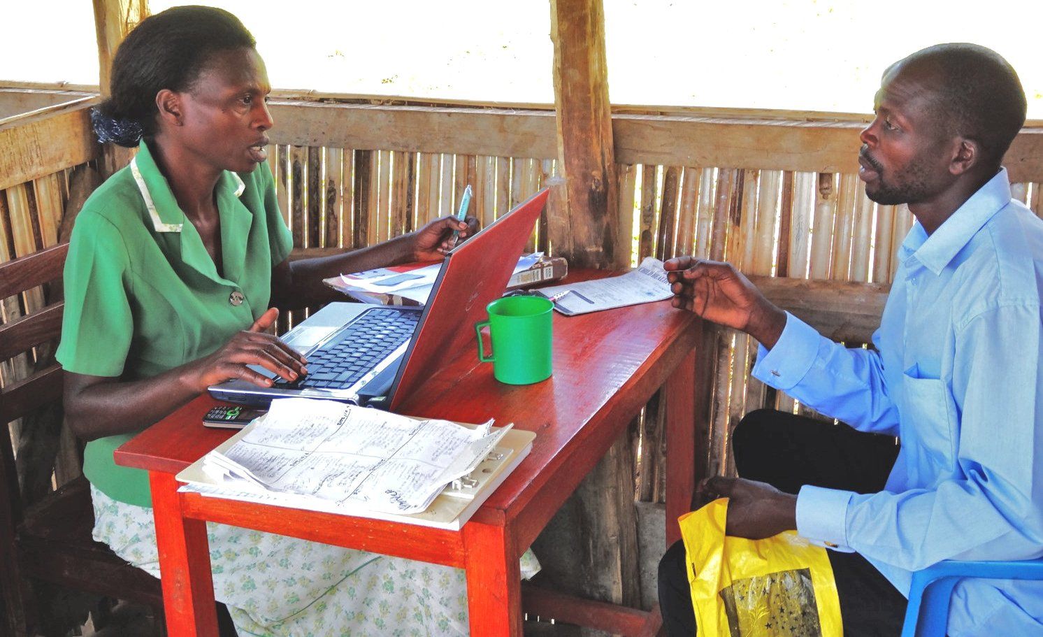 Woman using laptop, talking to a man. They are seated at a table outdoors under a shelter.