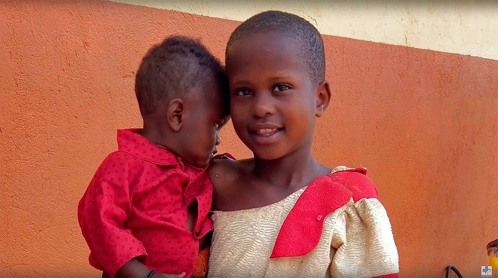 Girl in cream and red dress holding a child in a red shirt, smiling, against an orange wall.