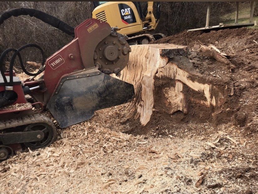 Excavator with stump grinder cutting a tree stump, throwing wood chips on a dirt site.