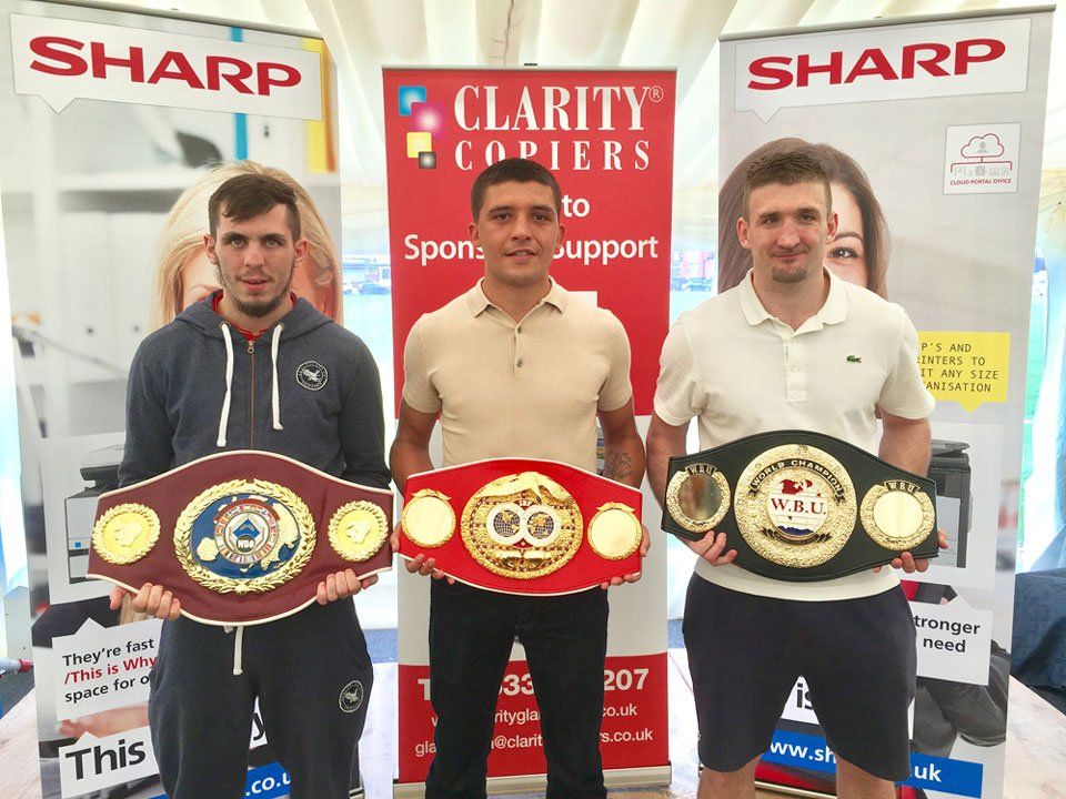 Three men holding boxing belts in front of a sharp sign