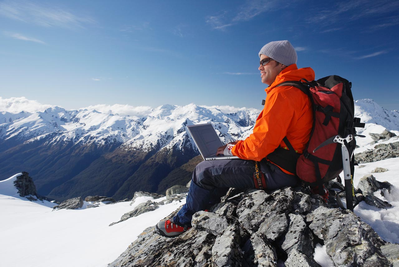 A man sits on top of a snow covered mountain using a laptop