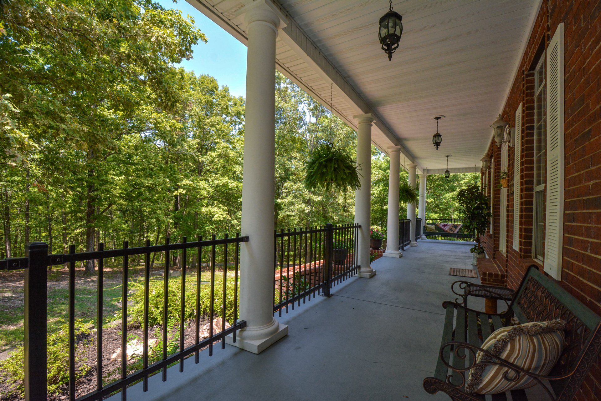 A long covered porch with white columns, black railing, and brick wall, overlooking a wooded area.
