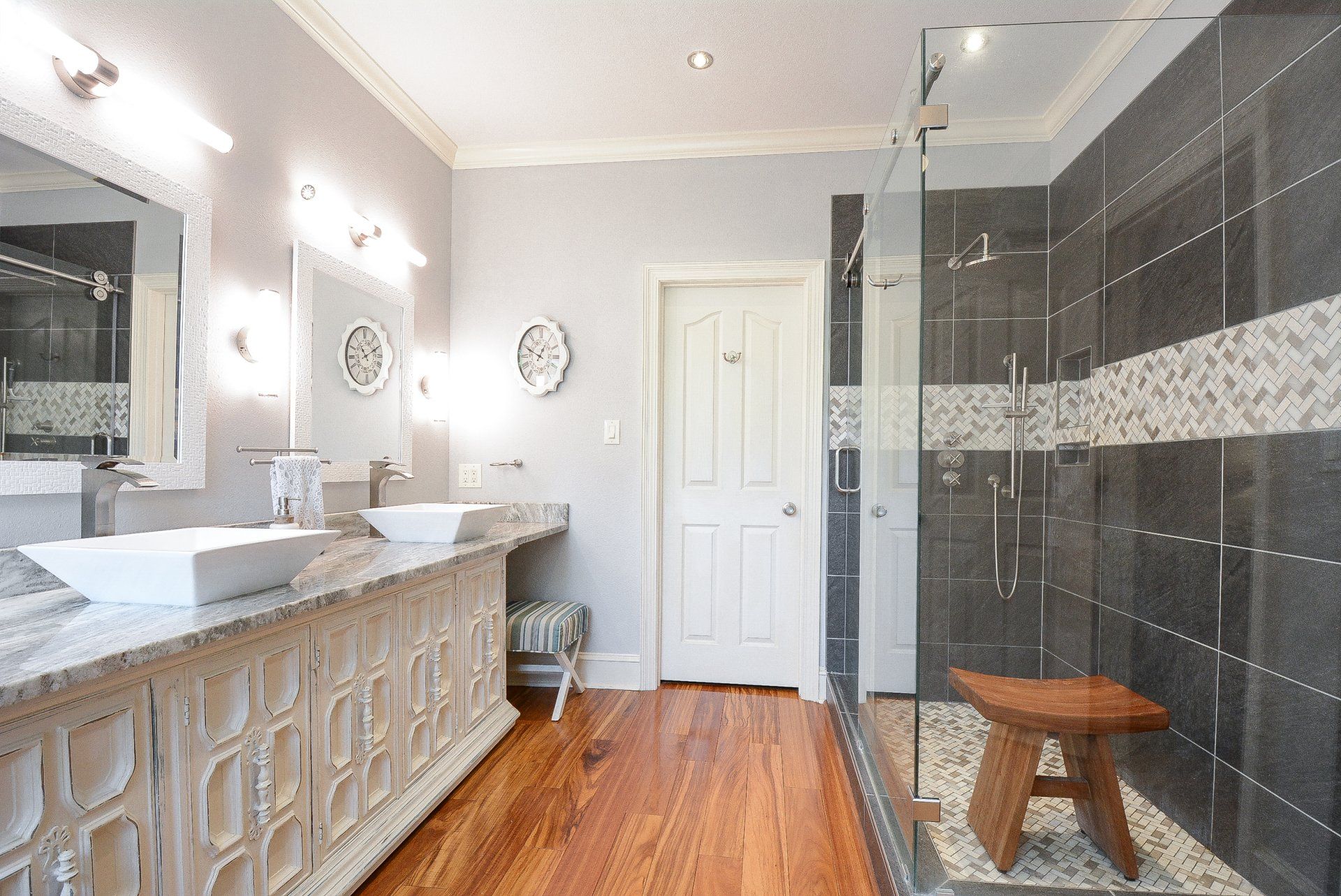 Bathroom with dual vanity, walk-in shower, wooden stool, and hardwood floor.