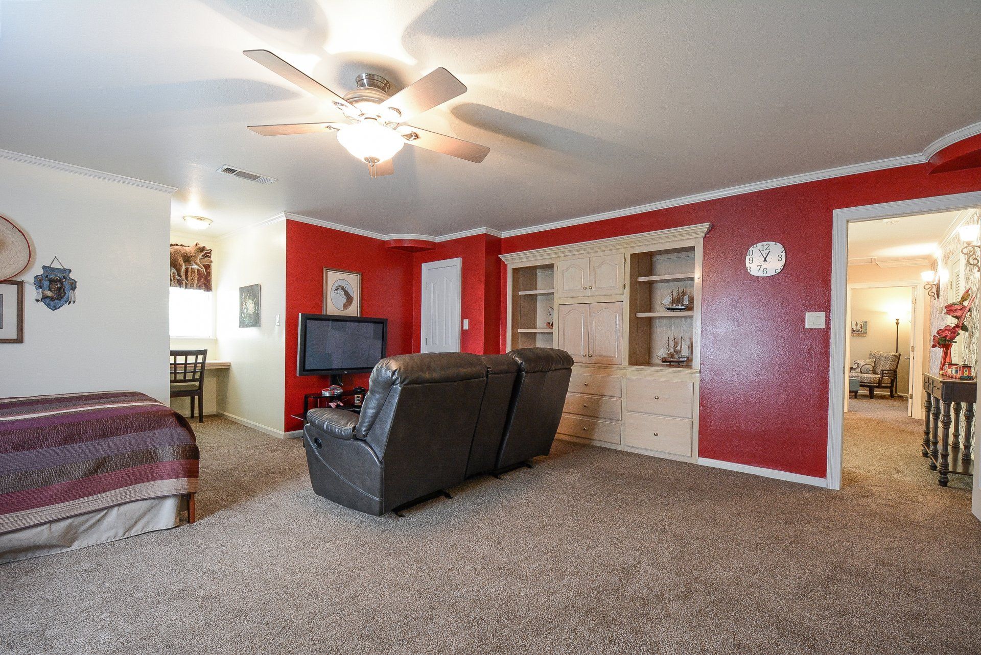 Living room with red walls, brown carpet, and a reclining sofa; a bed is visible in the background.