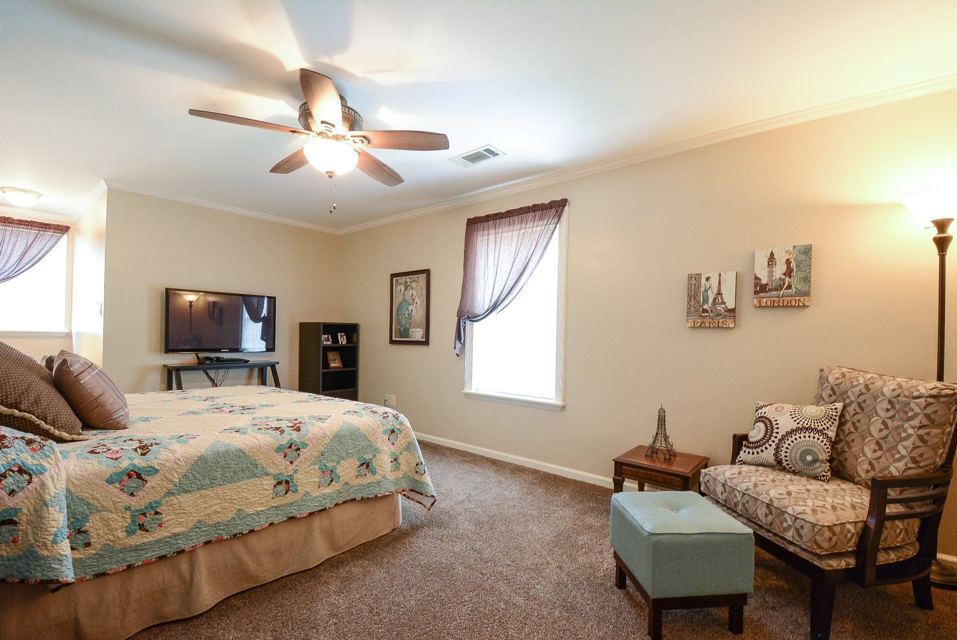 Bedroom with bed, TV, chair, and window. Beige walls, brown carpet, floral bedding, and patterned chair.