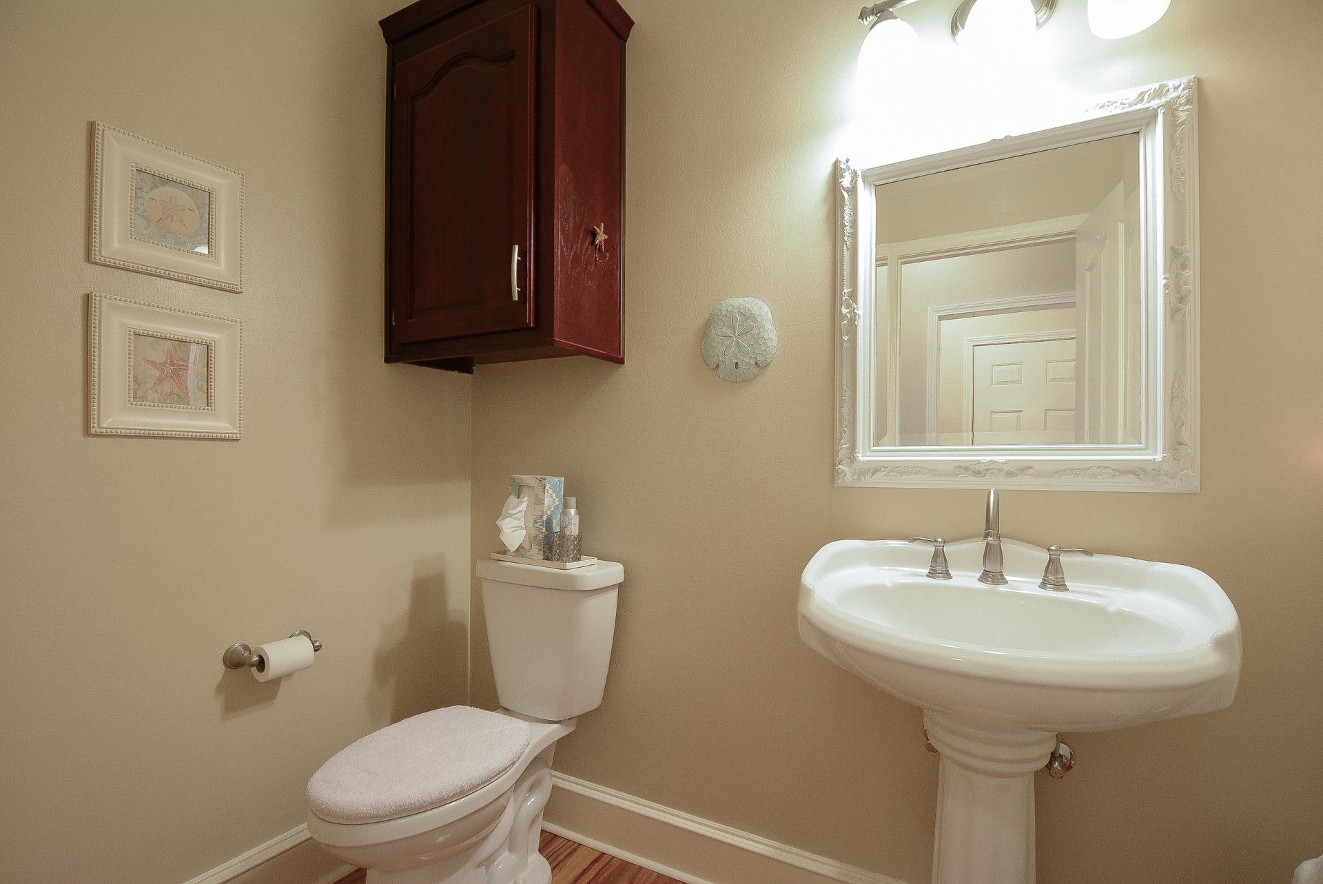 A small bathroom with a toilet, sink, mirror, and wall-mounted cabinet, cream walls, and a dark wood door.