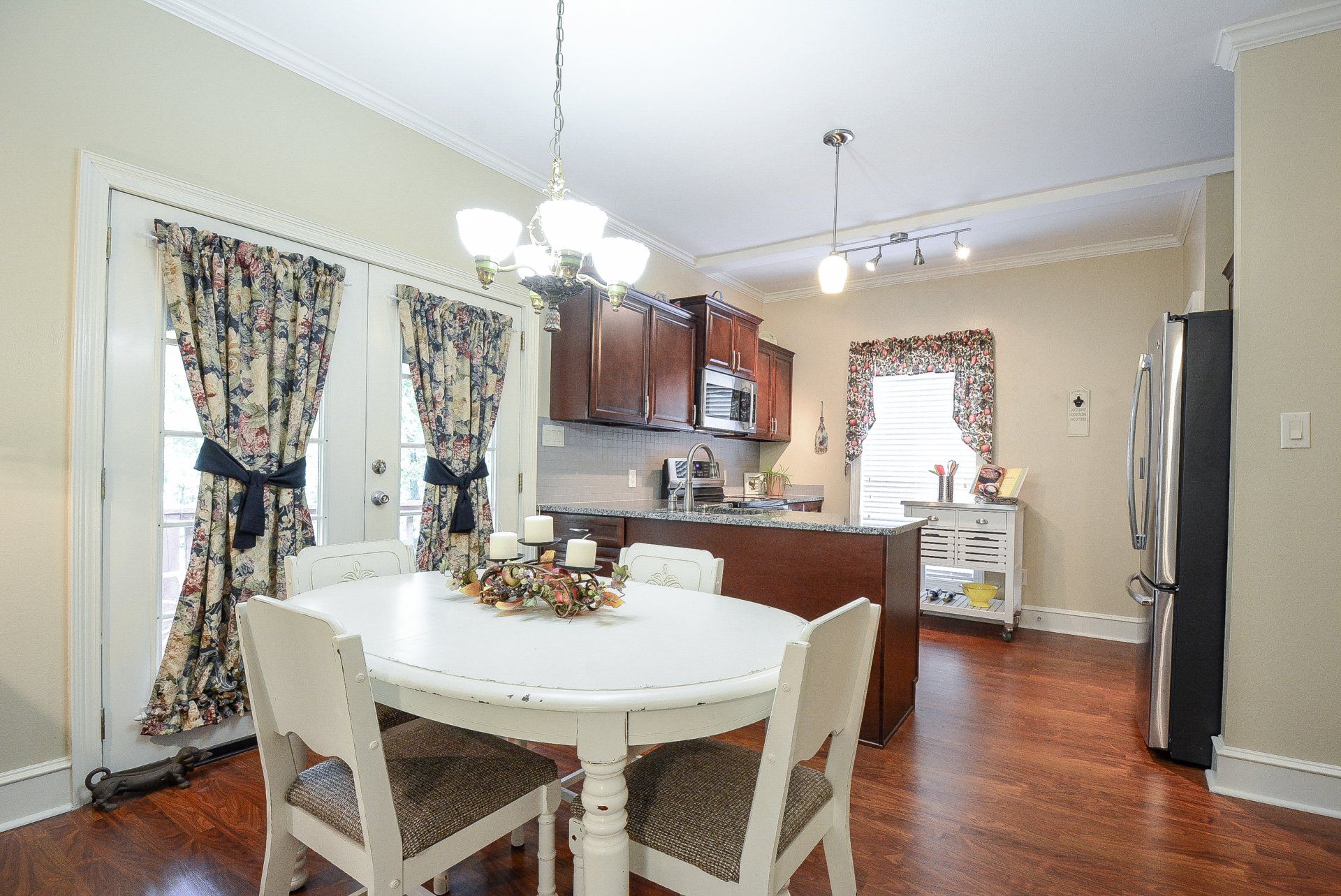 Dining area with white table, four chairs, and floral curtains; kitchen in the background.