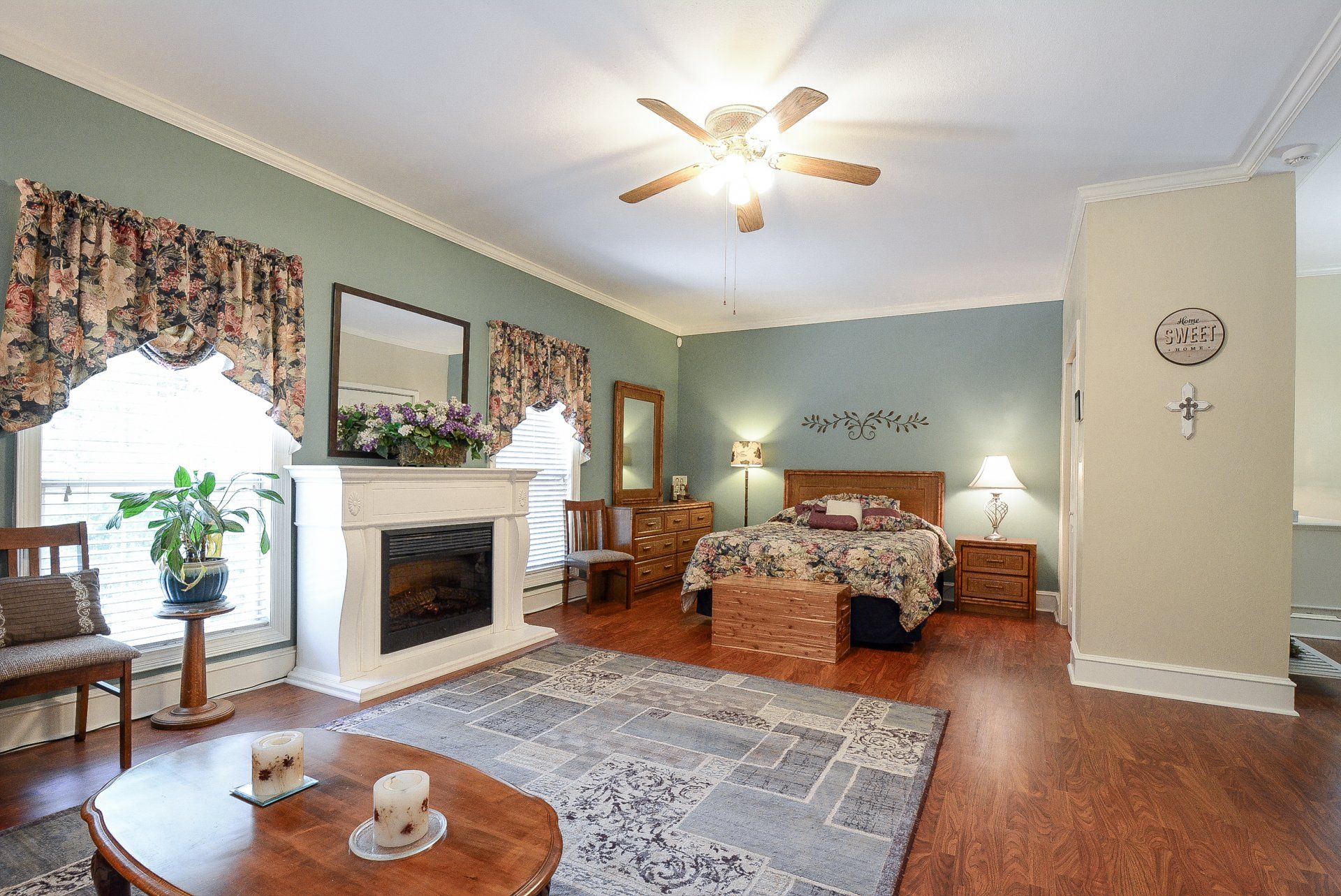 Bedroom with fireplace, bed, furniture; blue walls, patterned curtains, wooden floors, ceiling fan.