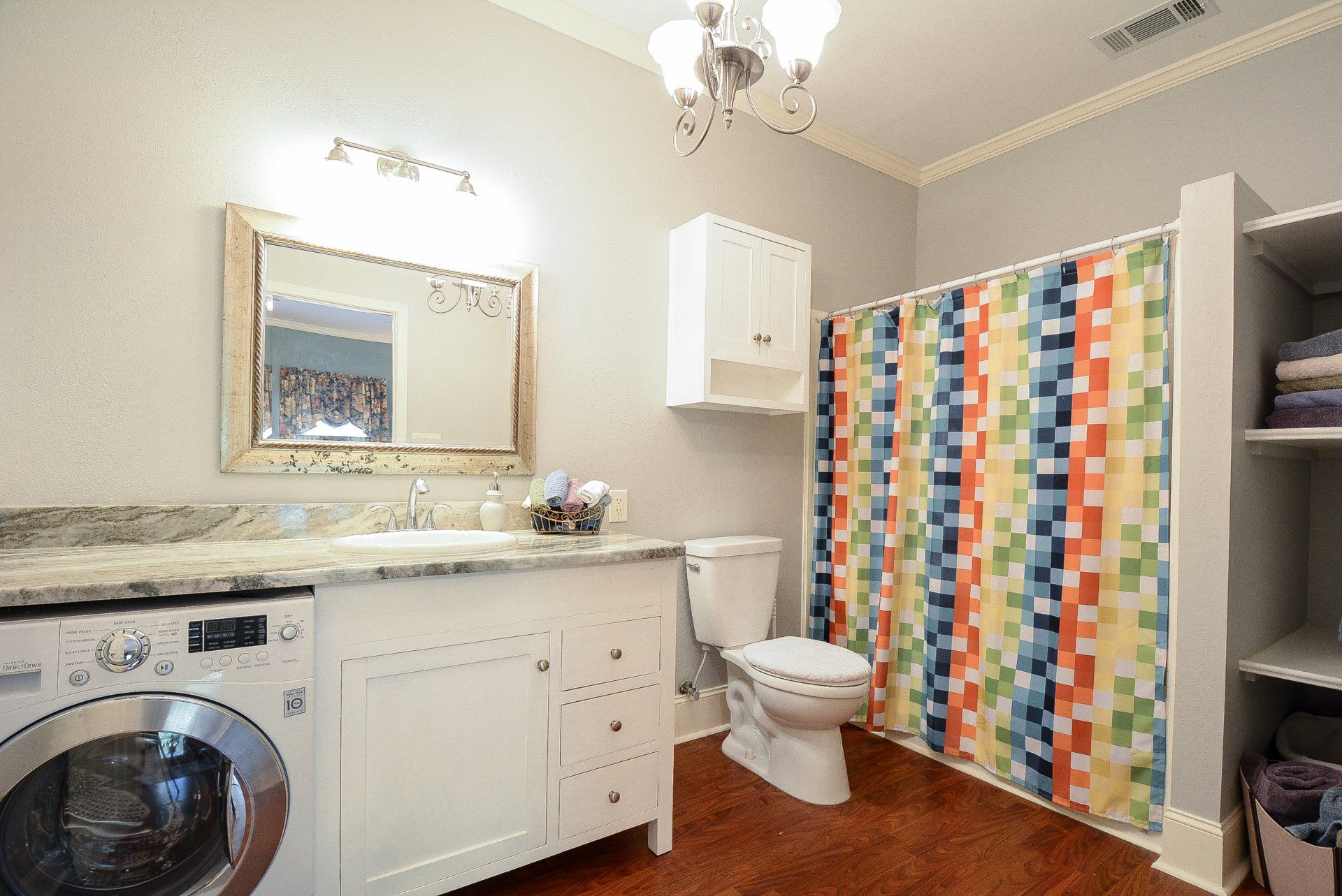 Bathroom with washing machine, vanity, toilet, shower with colorful curtain, and shelving.