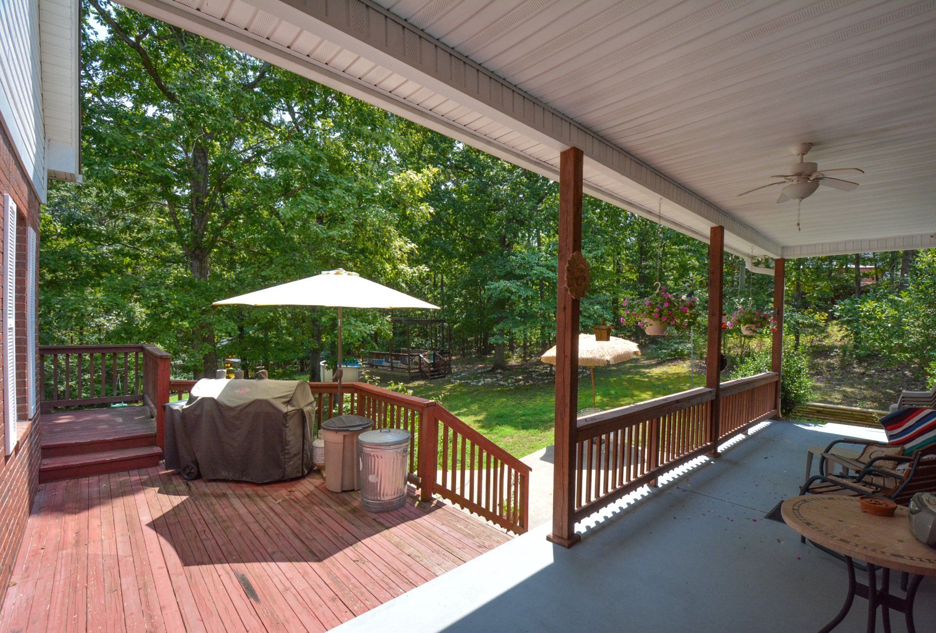 Covered porch with a wooden deck, overlooking a green yard with trees. A patio table and chairs are on the right.