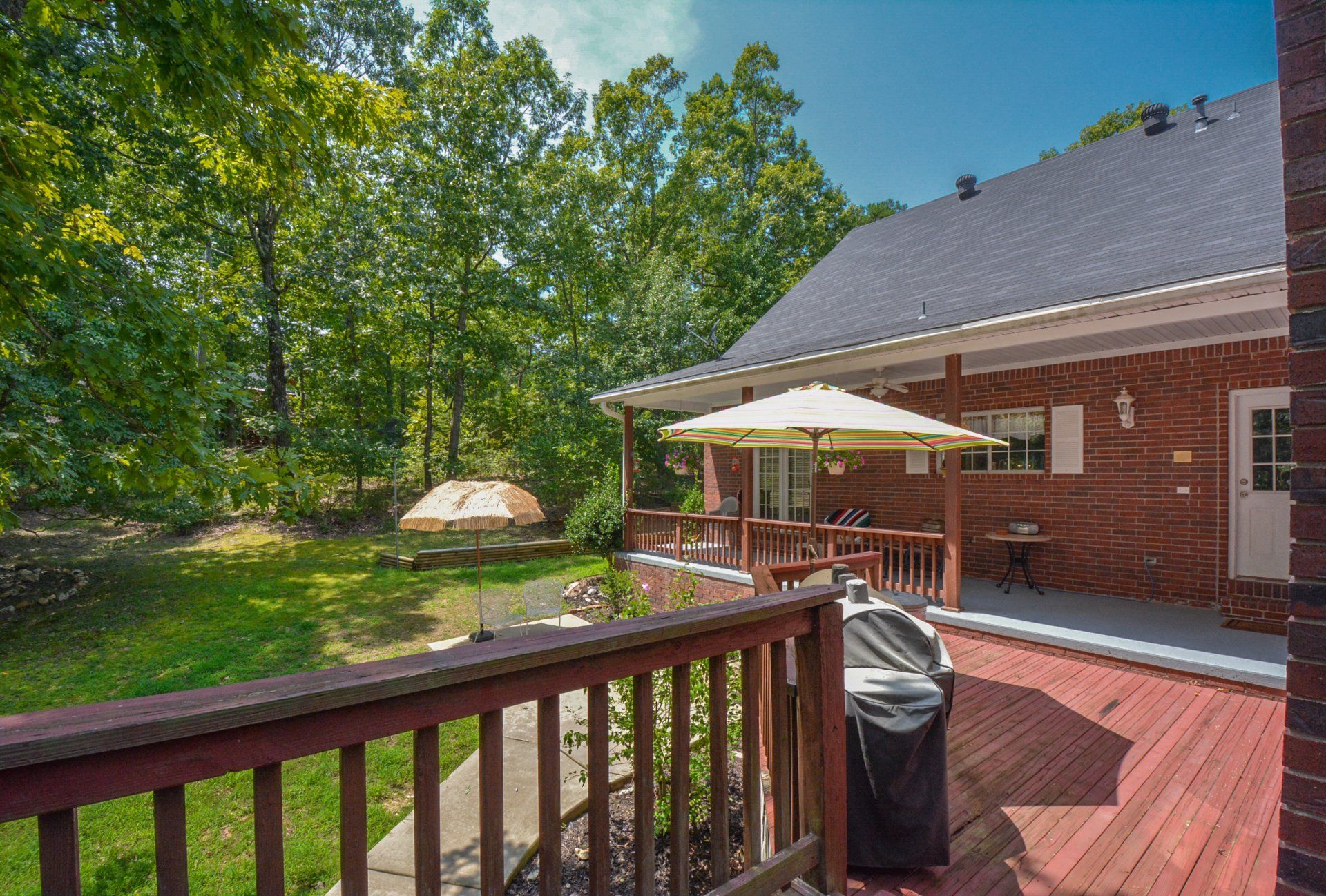 Wooden deck with railing, brick house, grill, umbrella, and backyard trees.