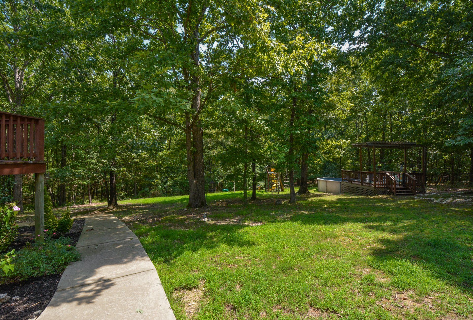 Grassy backyard with trees, a concrete path, and a deck in a wooded area.