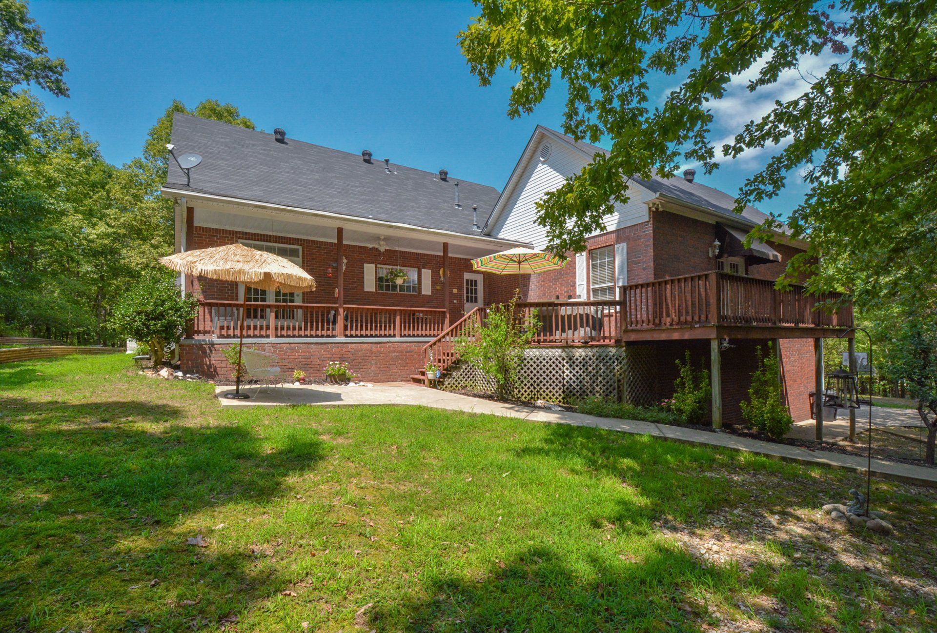 Brick house with a wraparound porch and deck, nestled in a green, tree-filled yard under a blue sky.