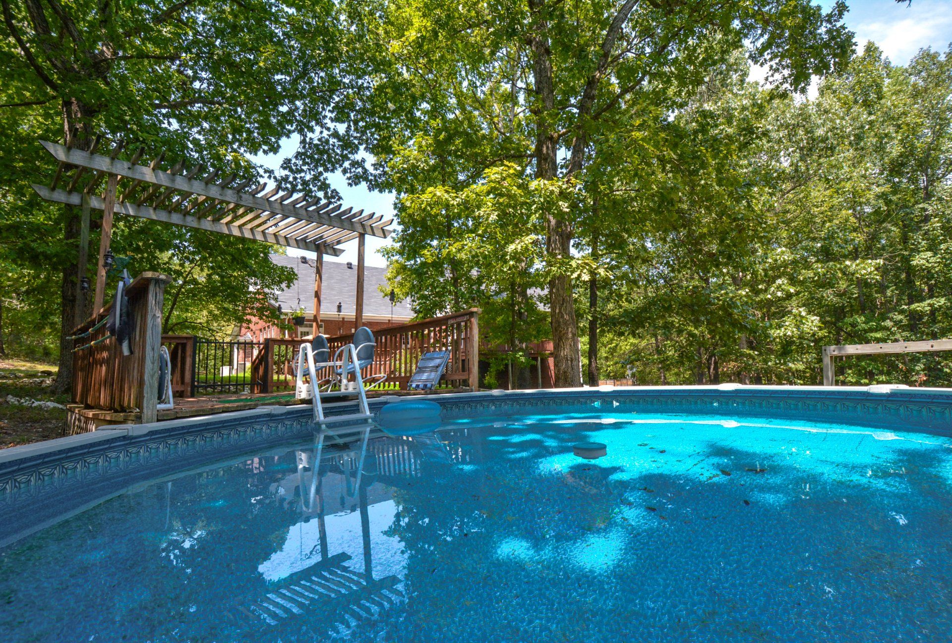 Swimming pool with ladder, surrounded by trees and wooden deck. Blue water, sunny day.