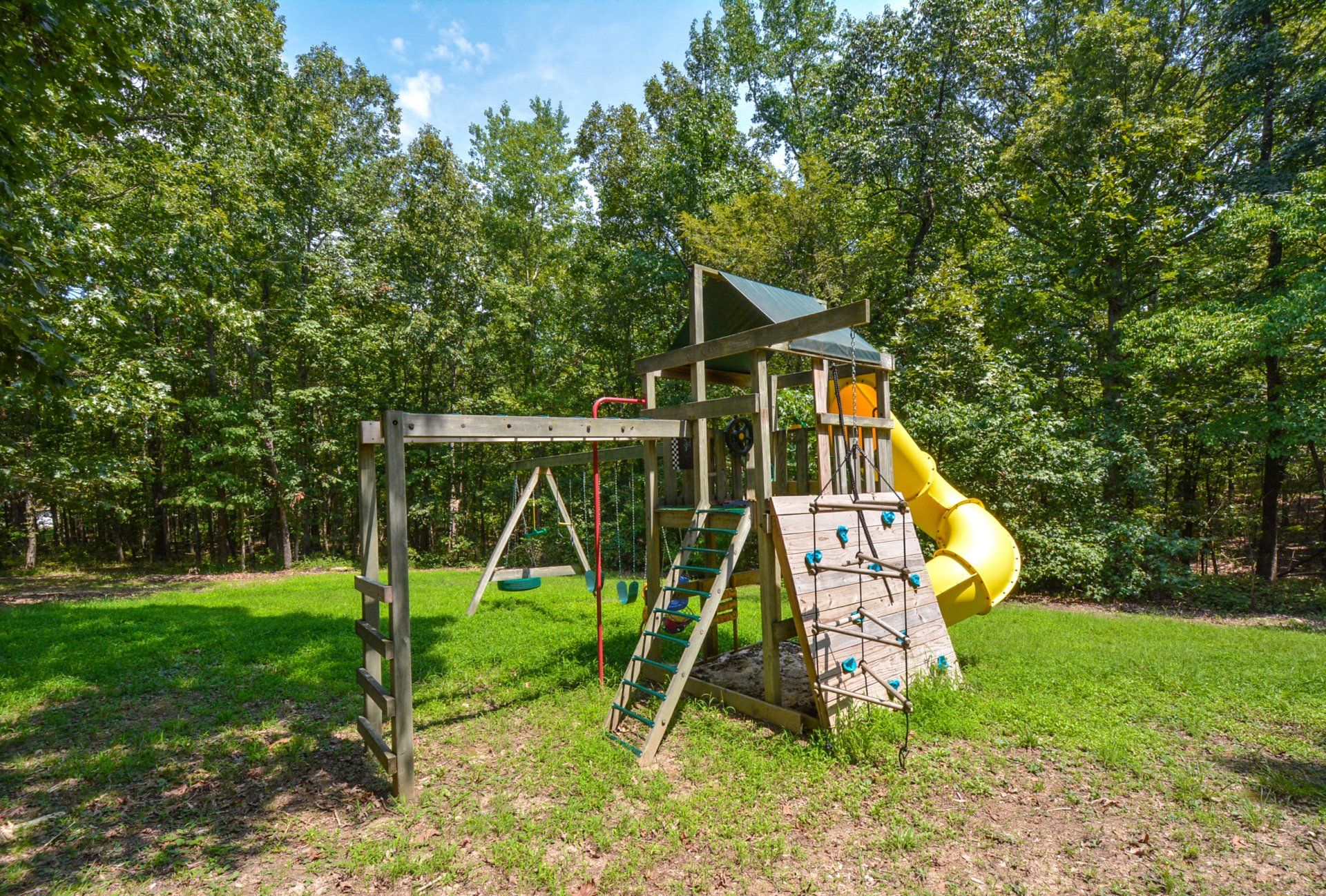 Wooden playground set in grassy yard, with slide, swings, climbing wall, and surrounding trees.