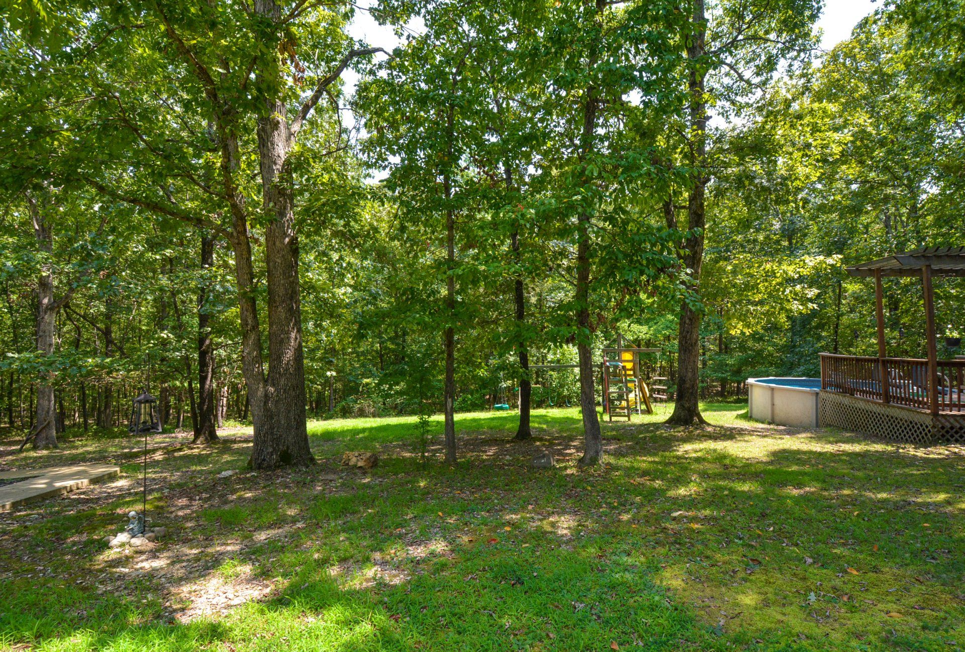 Lush backyard with trees, a swing set, small pool, and a wooden gazebo on a sunny day.