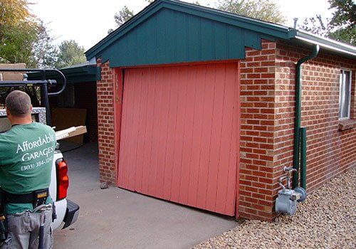Man in green shirt near pink garage door, brick building, daylight.
