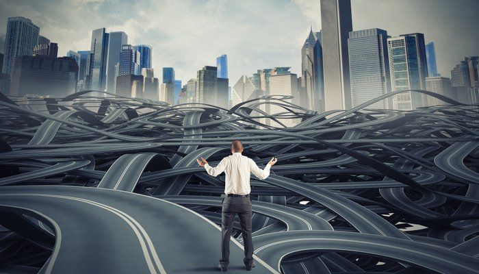 Man in business clothes, facing a skyline, looks toward multiple winding highways and shrugs.