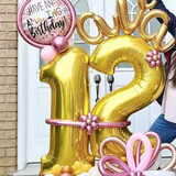 A woman is standing in front of a door with balloons in the shape of the number 12.