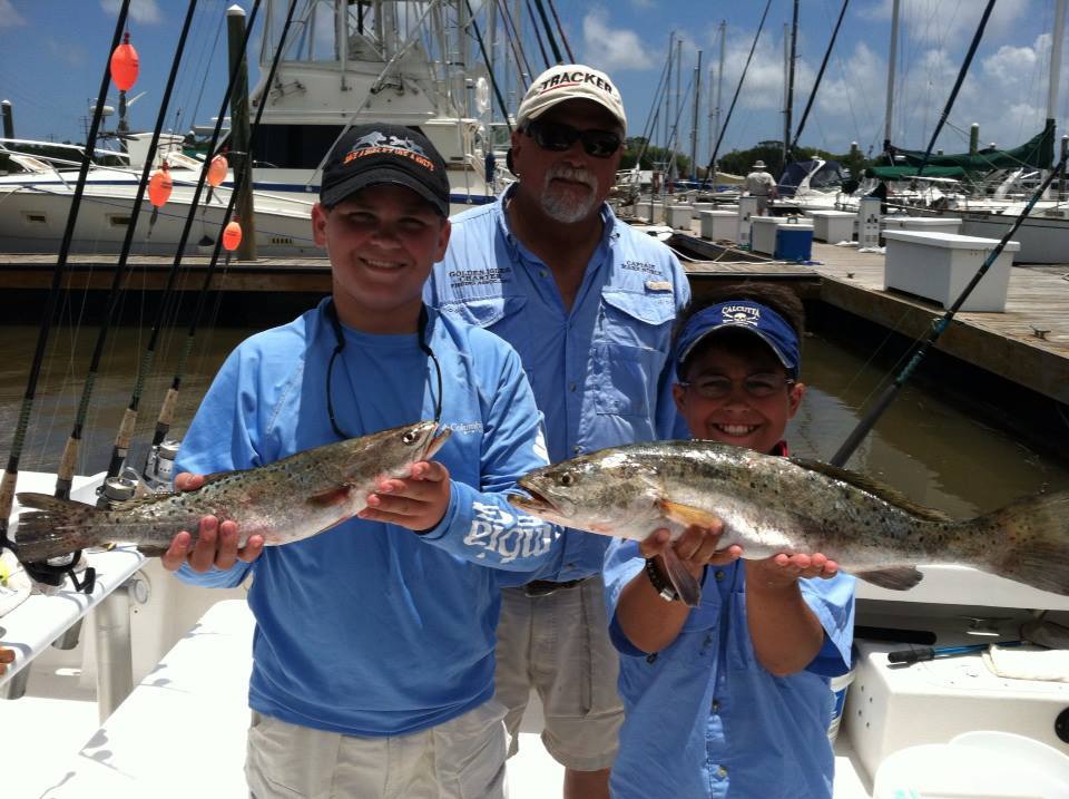 Two boys and an adult man on a boat, holding up caught fish at a dock on a sunny day.