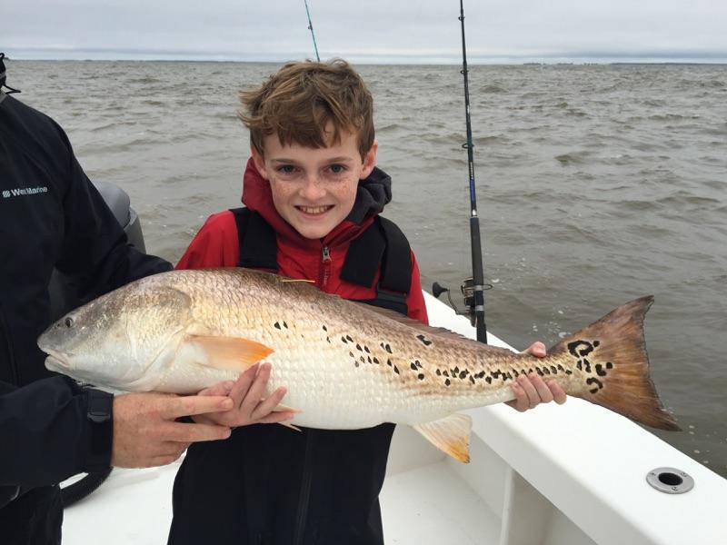 Boy smiling, holding a large redfish on a boat, overcast day.