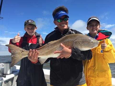 Person and two kids on a boat, holding up large redfish, giving thumbs up.