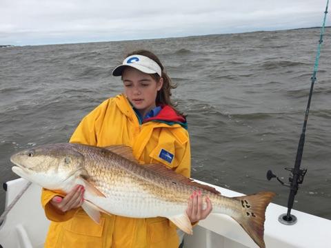 Girl in yellow raincoat holds large redfish on a boat, overcast day.