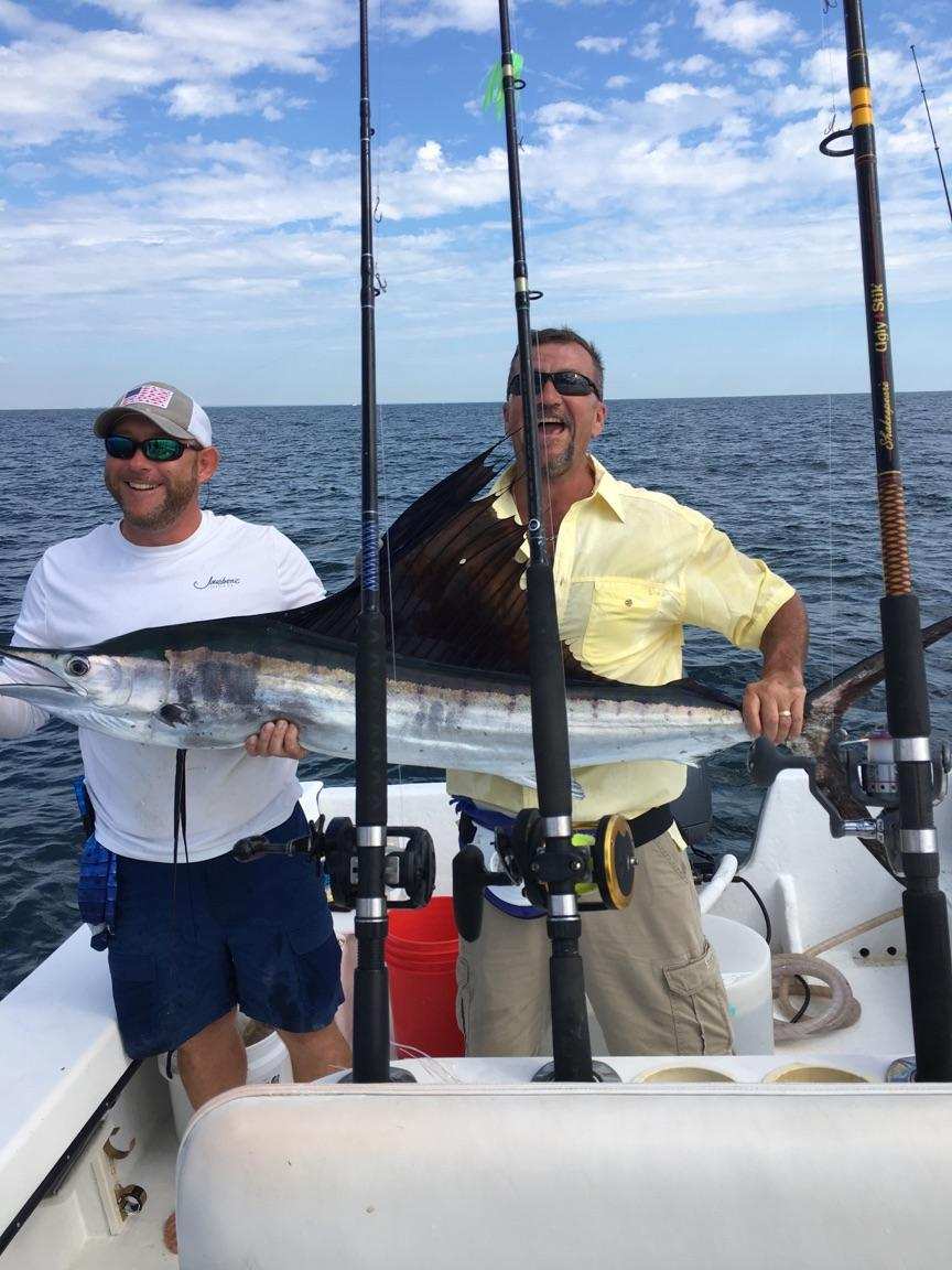 Two men on a boat, holding a large sailfish. Ocean in background, sunny day.