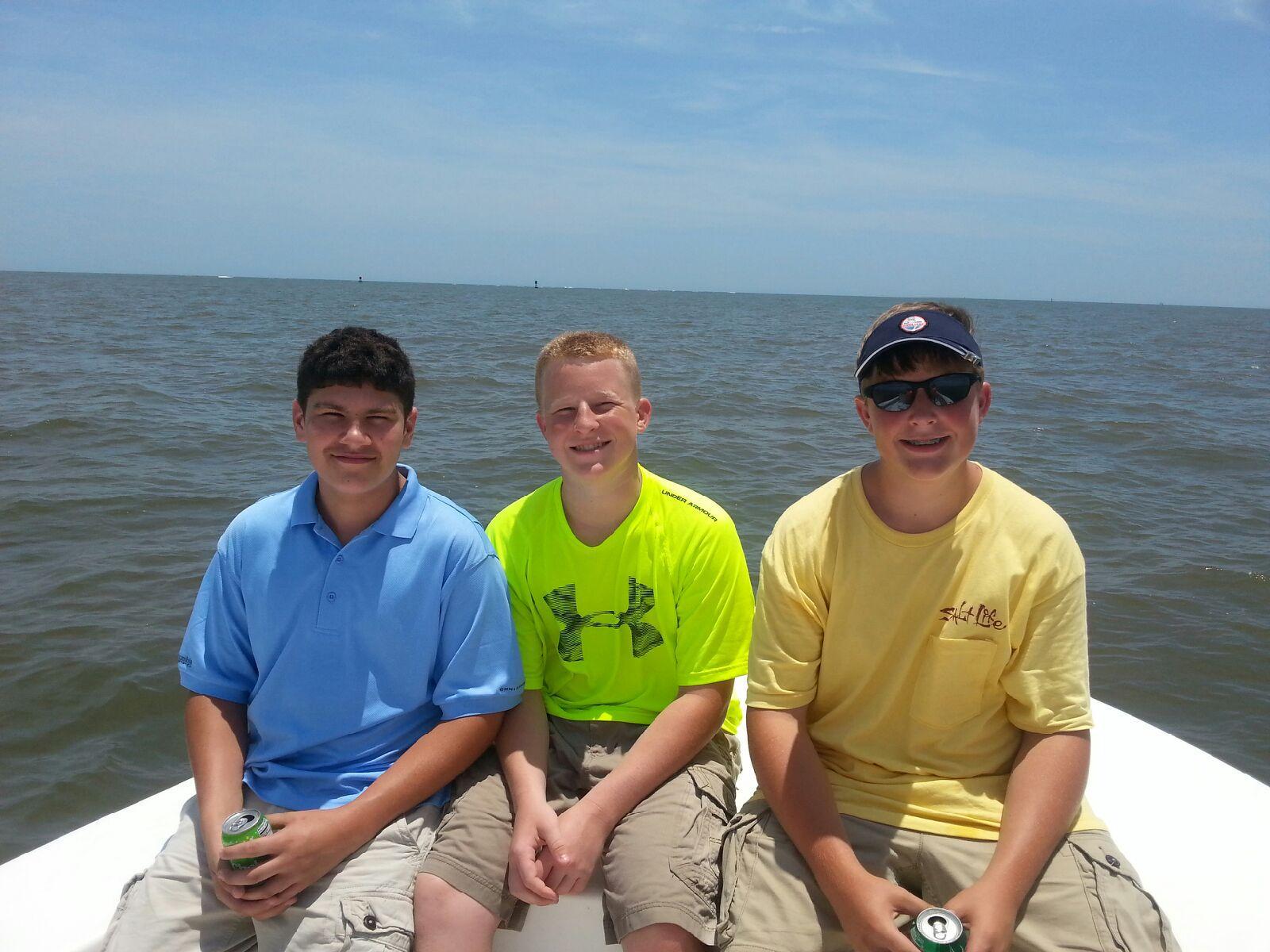 Three boys on a boat, smiling, with a blue sky and water background.