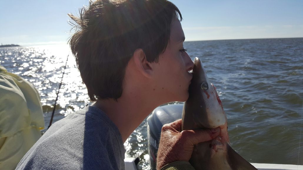 Teenager kissing a small shark on a boat with water in the background. Sunny day.