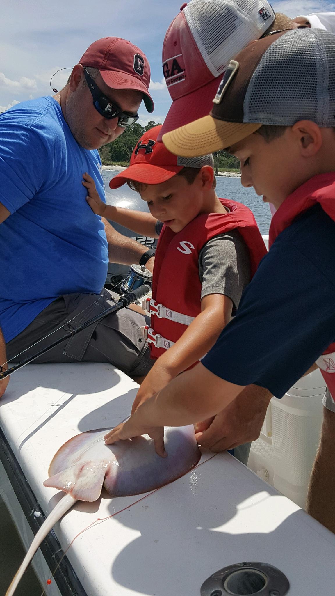 People on a boat examine a gray stingray. Two young boys in life vests touch it, with adults looking on.