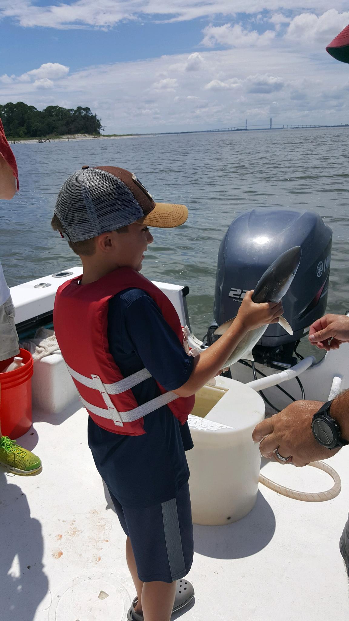 Boy on a boat smiles, holding a fish. The sky is blue, and the water is visible in the background.