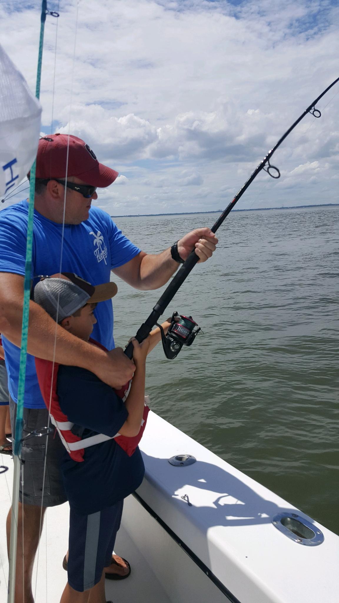 Man assists child holding fishing rod on a boat, blue water and sky.