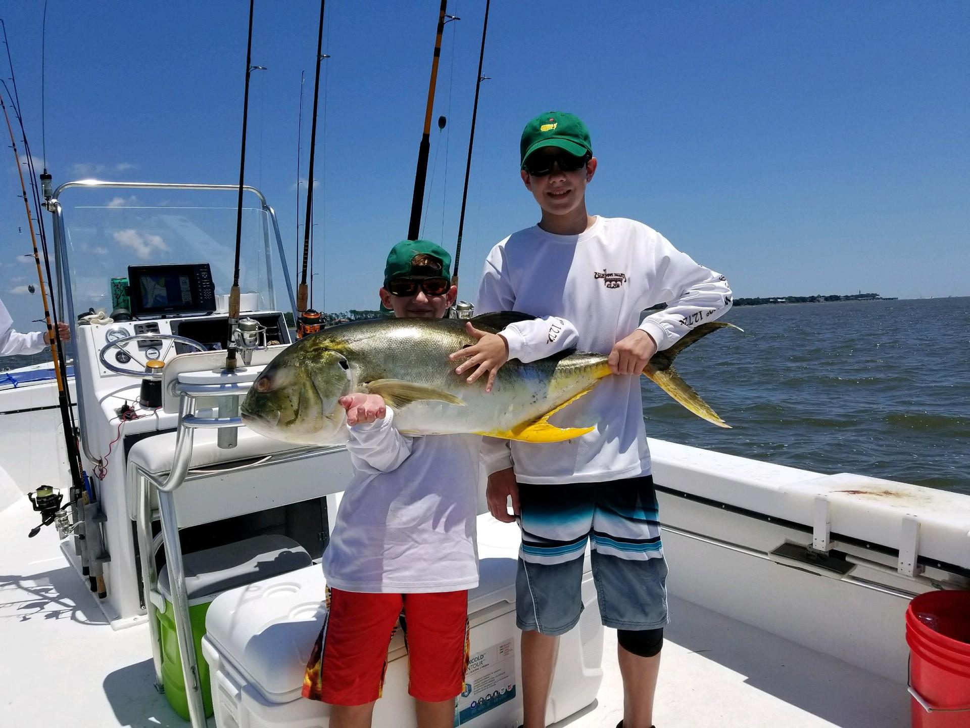 Two boys holding a large yellow jack fish on a boat, sunny day.